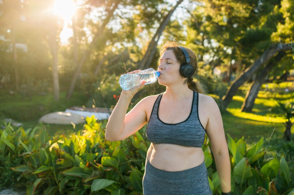 Overweight woman drinking water after jogging in the park. Portrait of young plus-size thirsty woman