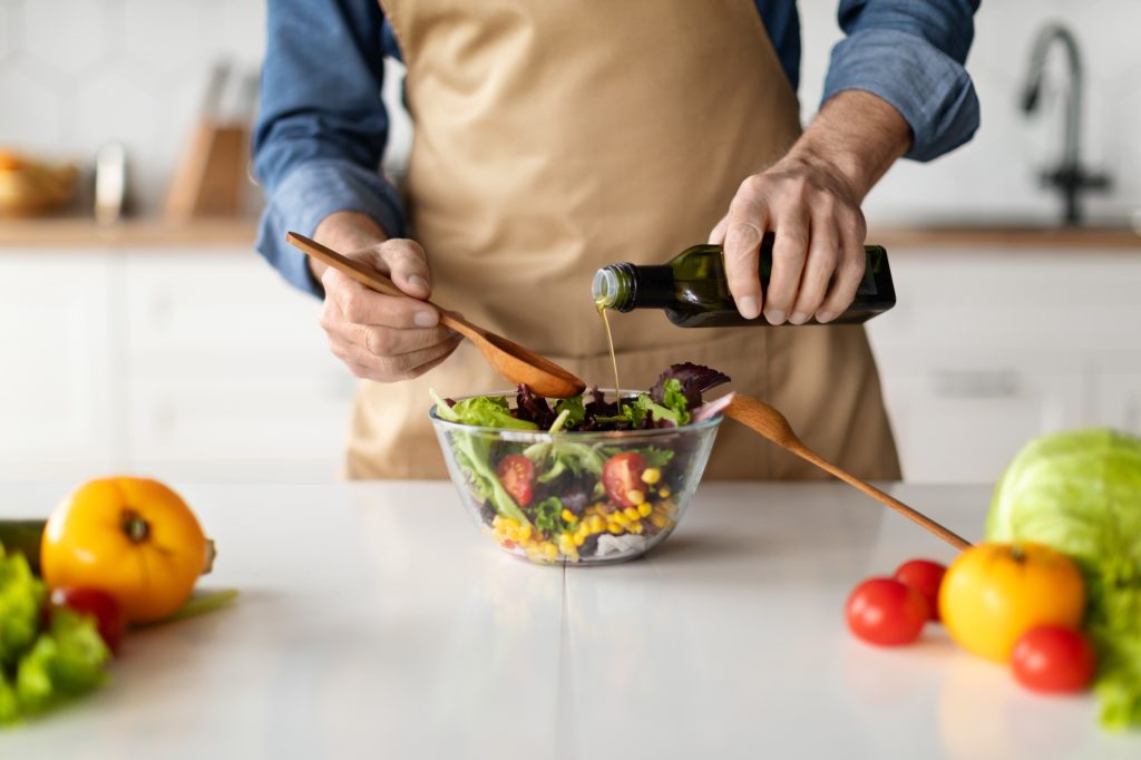Healthy Food Concept. Mature Man Seasoning Vegetable Salad While Cooking In Kitchen